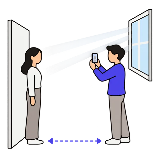 One person standing in front of a plain wall while another holds a phone at eye level a few feet away, with light from a window